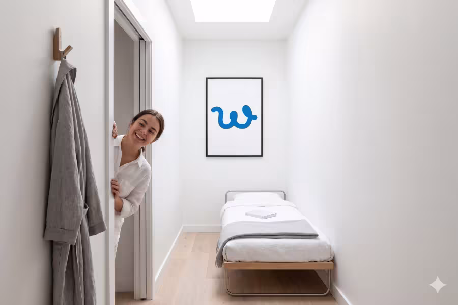 A humorous yet highly aesthetic minimal lifestyle shot of an impossibly narrow, pristine white room. A single, minimalist cot is neatly made.