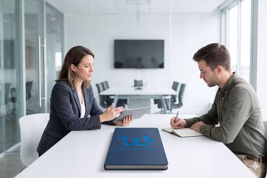 Minimal lifestyle office photography. A bright, modern glass-walled meeting space. Two people are sitting across from each other at a sleek white table, one acting as a consultant, the other taking notes.