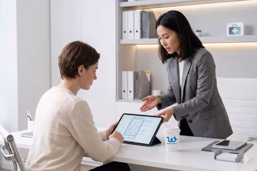 Minimalist lifestyle photography of a modern professional clinic office. A human client is seated at a sleek white desk, attentively viewing a large digital tablet displaying a clean wireframe graphic. A professional consultant stands nearby, gesturing toward the screen.